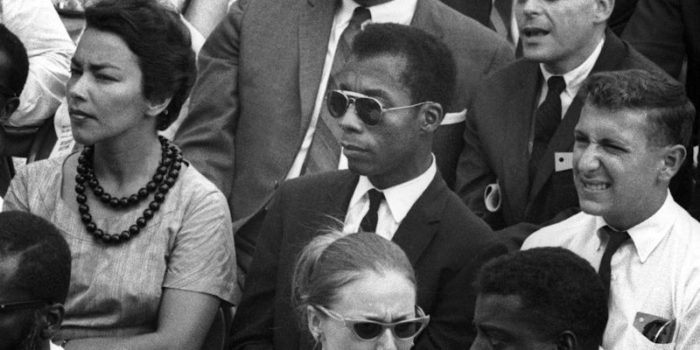 Monochrome photograph showing a youngish James Baldwin, in suit and sunglasses, sat in an audience. They seem to be sat outdoors. To Baldwin's left is a white man in a suit squinting from the sun and smiling, to his right is a woman in a short-sleeved dress and bead necklace with short hair. You can also see two Black men and a white woman (in sunglasses) in the row in front of him, and a white man behind.